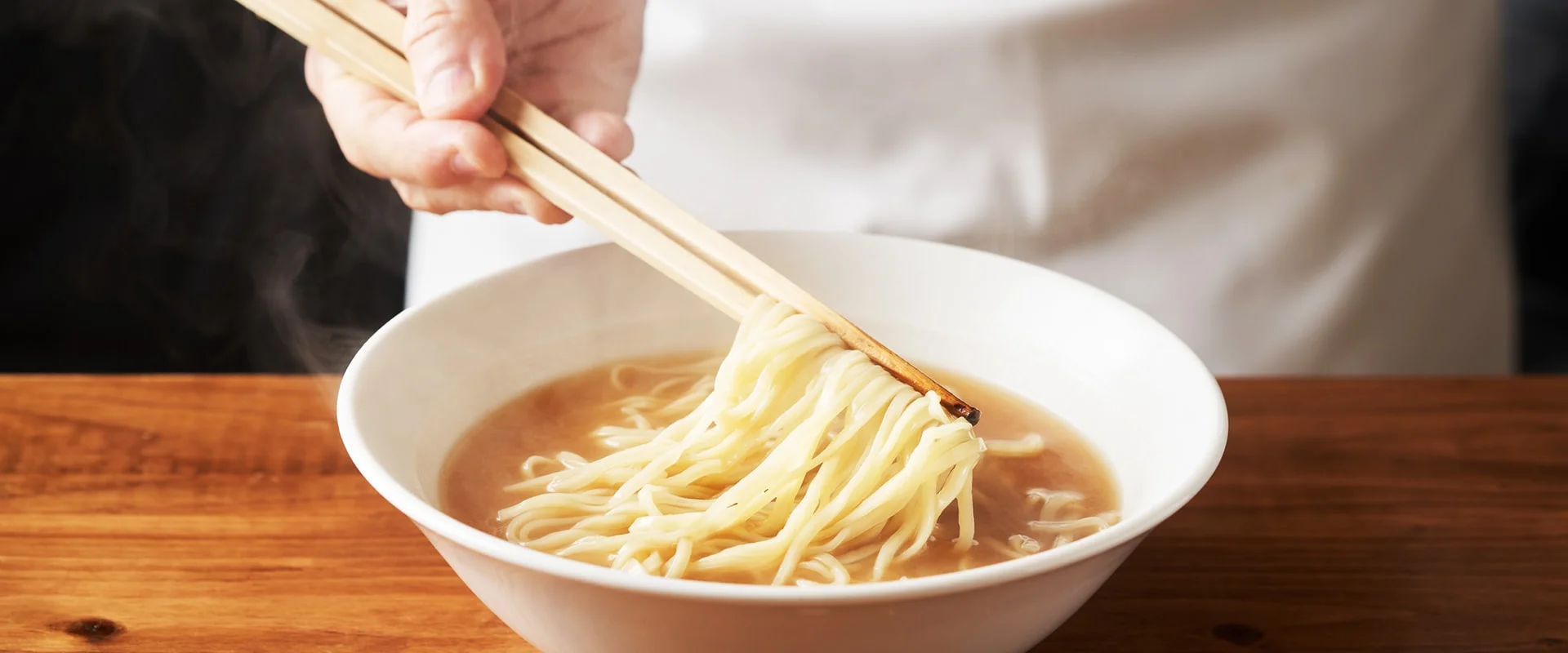 箸で持ち上げたつやのある中華麺と湯気の立つラーメンの丼